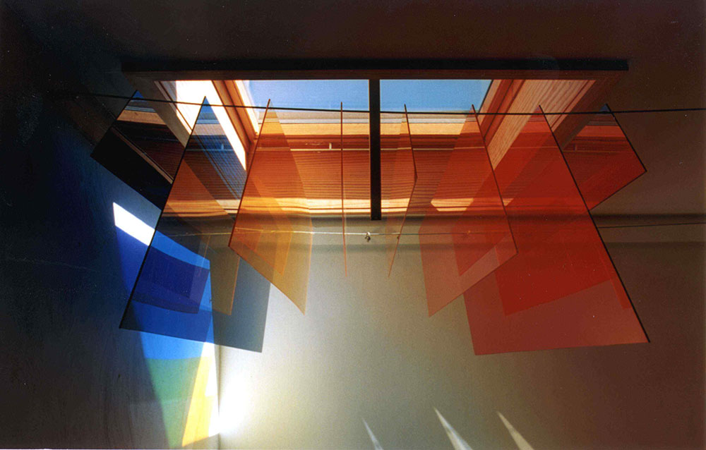 Ein Kunstwerk im öffentlichen Treppenhaus. Es besteht aus transparenten farbige Scheiben, in den Farben Blau, Gelb, Orange und Rot. Sie hängen in unterschiedlicher Länge unter dem Dachfenster. Durch das Sonnenlicht sind farbige Lichtreflexe an den Wänden zu sehen. Das Farbspiel ist zur Sommersonnenwende aufgenommen.
