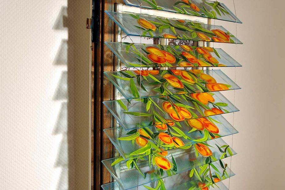 In front of an old wooden window, mounted on the wall, there is a blind made of colored melted glass. The glass slats show peach fruits and branches of a tree. View from the side.