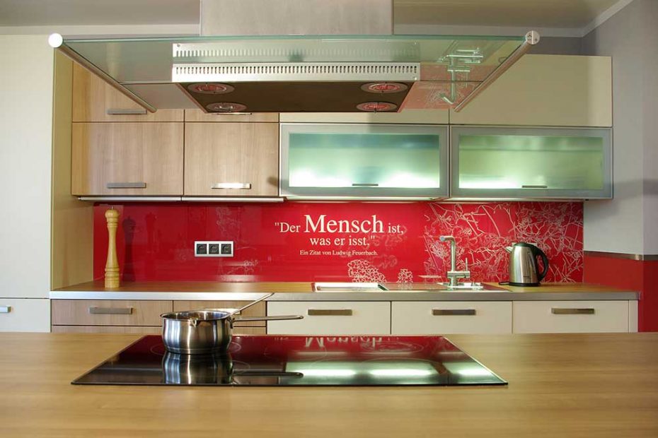 A kitchen with beige cabinets and wood units. The glass backsplash is printed with a pattern of white grass on a red background and a quote " You are, what you eat"