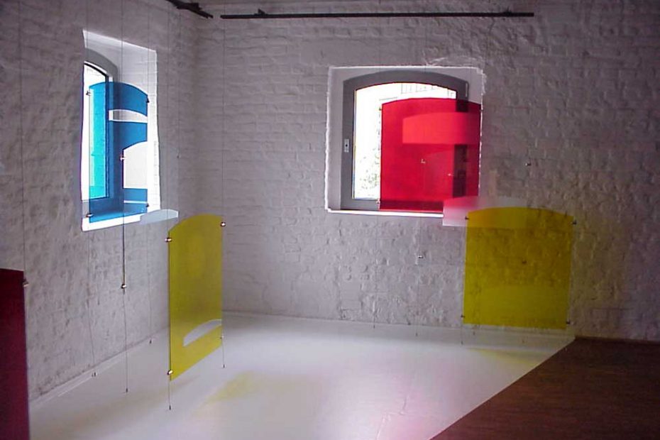In a white-painted brick room, 3 colored glass panels hang in front of 2 windows. They have a cutout or glued-on areas that mark the light reflections through the windows while the position of the sun changes.