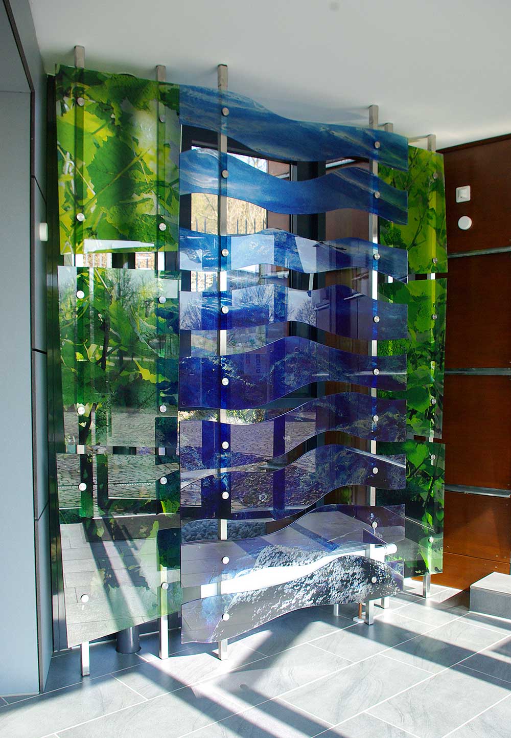 Art and public glass. Freely mounted glass panels attached to a stainless-steel frame in front of a window front. The panels on the right and left represent wine leaves. The curved panels in the middle represent the flowing water of the river Rhein. They are printed transparently and play with the light of the window front.