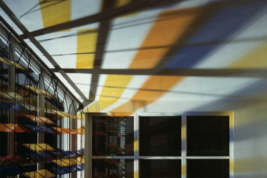 Due to a large, colored, transparent blind with sunlight from outside, light reflections in yellow, orange and blue appear on the ceiling of the room.
