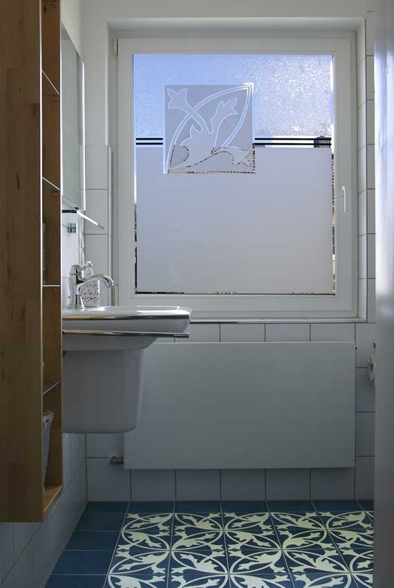 A bathroom window with a design that reflects the pattern of the floor tiles. The glass is partly sandblasted or has frosted ice flower pattern.